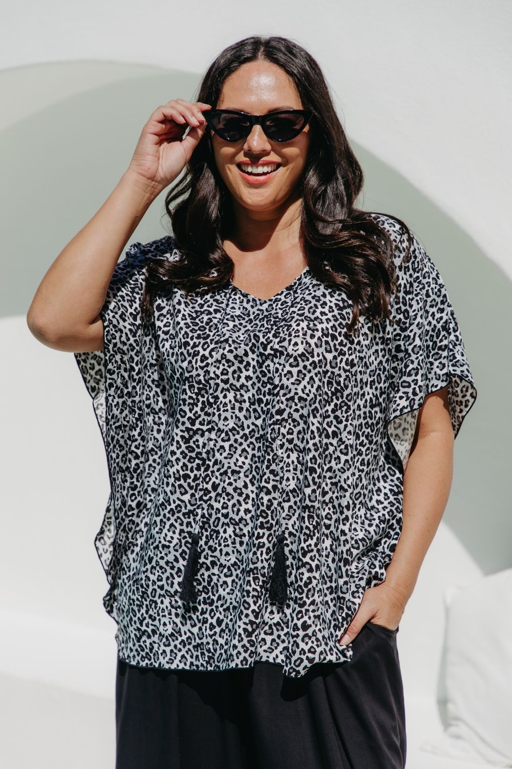 A woman wearing a black grey and white leopard print kimono sleeve kaftan top and black palazzo pants, posing outdoors against a white wall with a green arch.