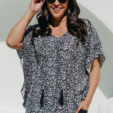 A woman wearing a black grey and white leopard print kimono sleeve kaftan top and black palazzo pants, posing outdoors against a white wall with a green arch.