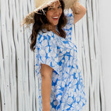 Model wearing a blue and white Hawaiian floral kaftan top with a large bamboo hat, looking back at the camera near a white bamboo fence