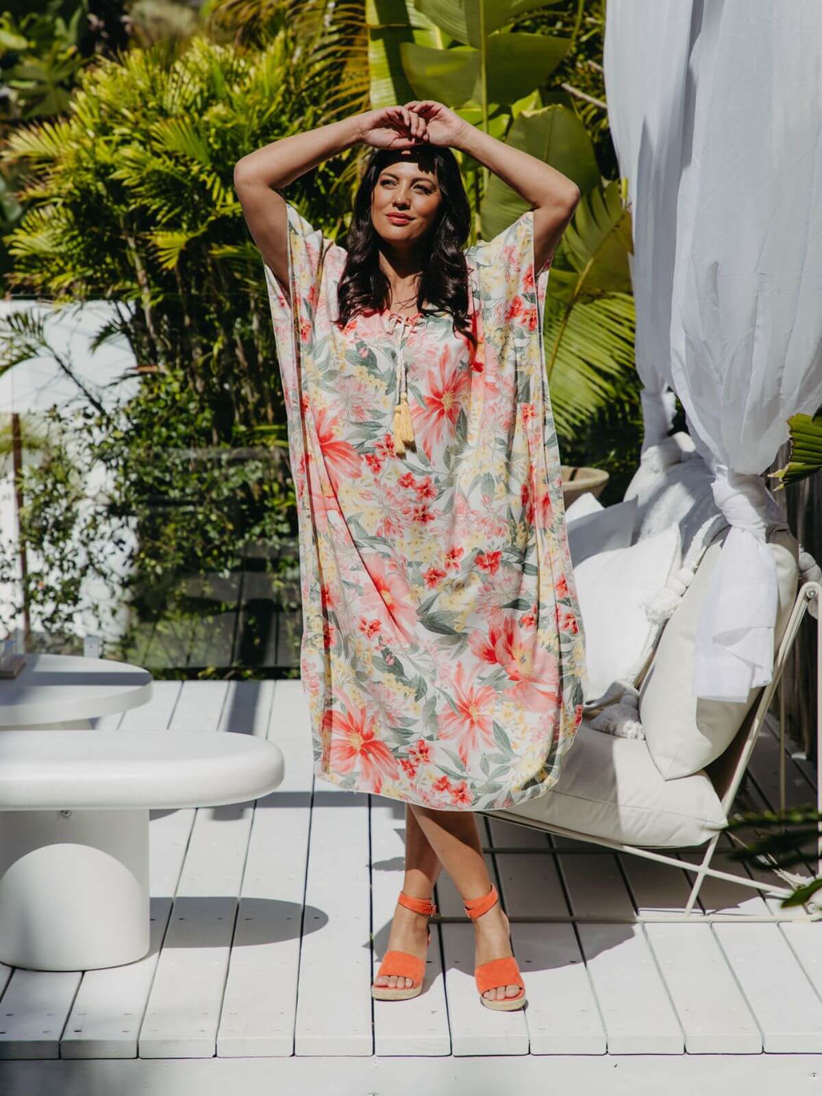 Model wearing a long floral kaftan with kimono sleeves standing on an outdoor deck with lounge seating