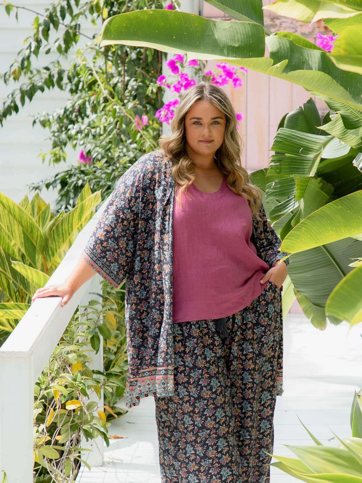 Plus size model wearing floral kimono jacket layered over grape linen top and matching pants in a tropical garden setting