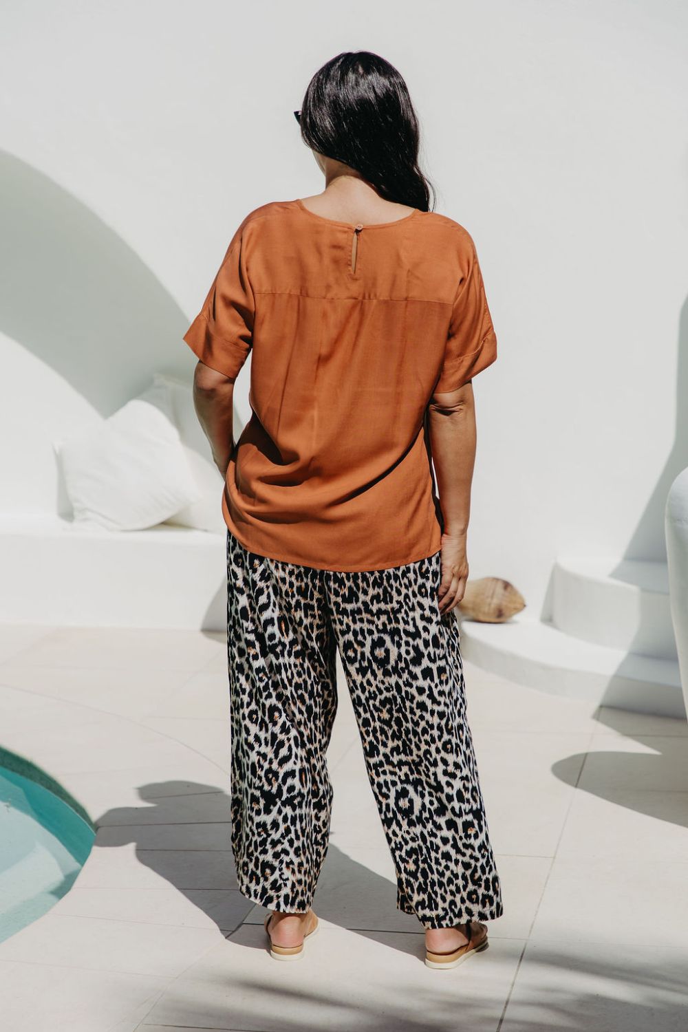 Lady wearing a caramel brown coloured top and leopard print pants standing in a sunlit area in the pool entertaining area