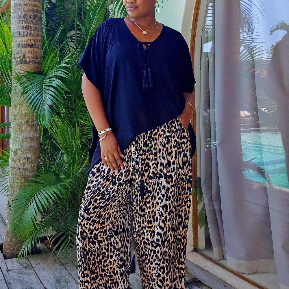 A woman stands barefoot on a wooden deck in her Bali villa wearing a black short-sleeve top and wide-leg leopard-print pants, with palm trees and a glass window in the background.
