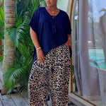 A woman stands barefoot on a wooden deck in her Bali villa wearing a black short-sleeve top and wide-leg leopard-print pants, with palm trees and a glass window in the background.