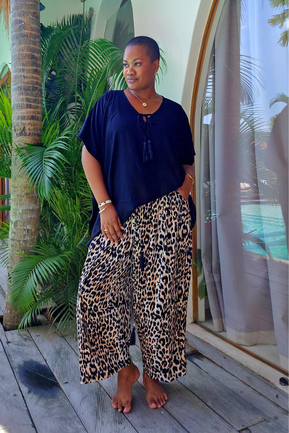 A woman stands barefoot on a wooden deck in her Bali villa wearing a black short-sleeve top and wide-leg leopard-print pants, with palm trees and a glass window in the background.
