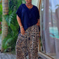 A woman stands barefoot on a wooden deck in her Bali villa wearing a black short-sleeve top and wide-leg leopard-print pants, with palm trees and a glass window in the background.