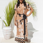 A woman models a kaftan maxi dress with a soft brass, black and soft white abstract print, featuring a black tie belt, standing in front of a white wall with a potted palm plant.