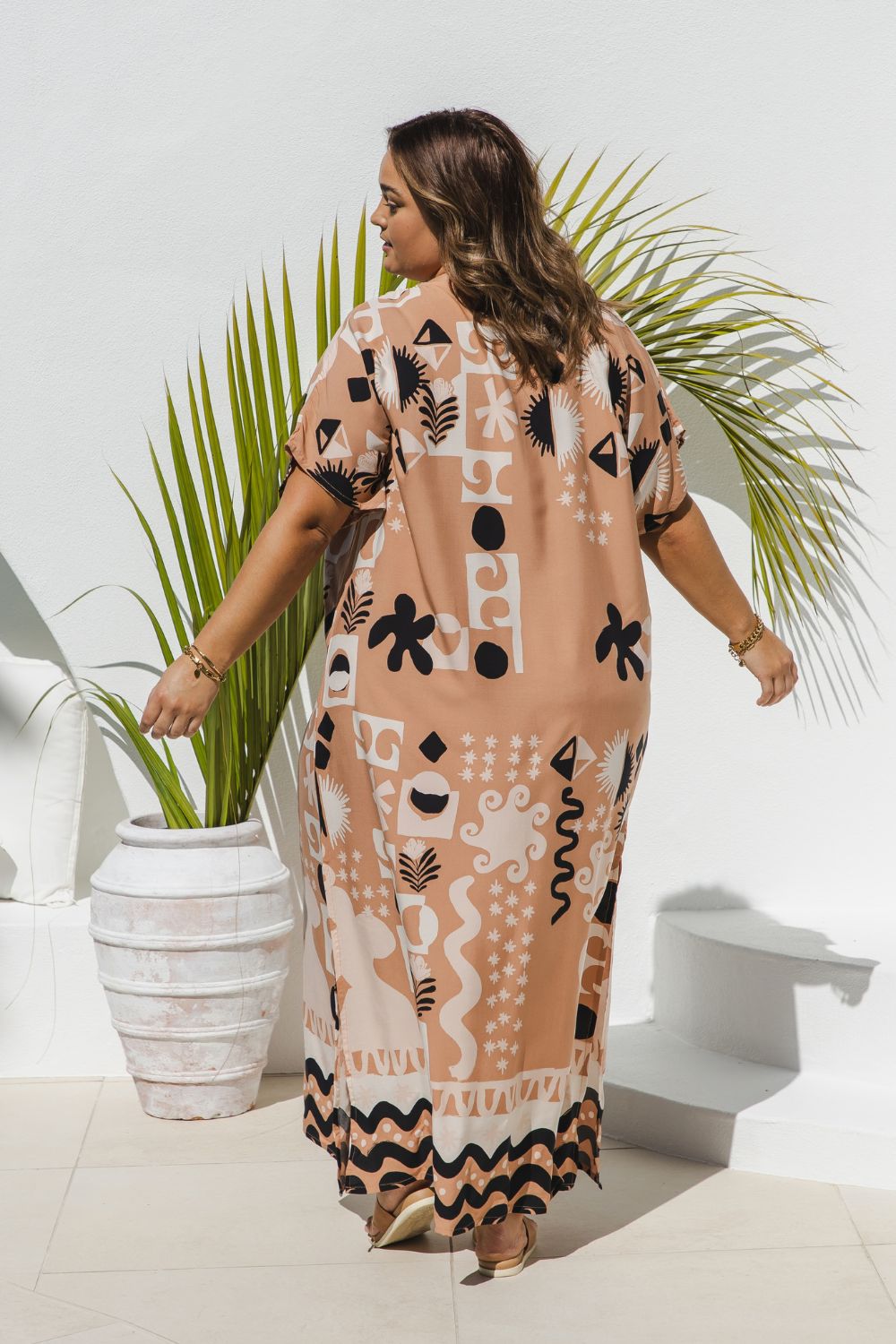 A plus size model wearing a long kaftan dress with black and white geometric patterns on soft terracotta, photographed from behind in a bright, modern outdoor setting with a potted palm plant.