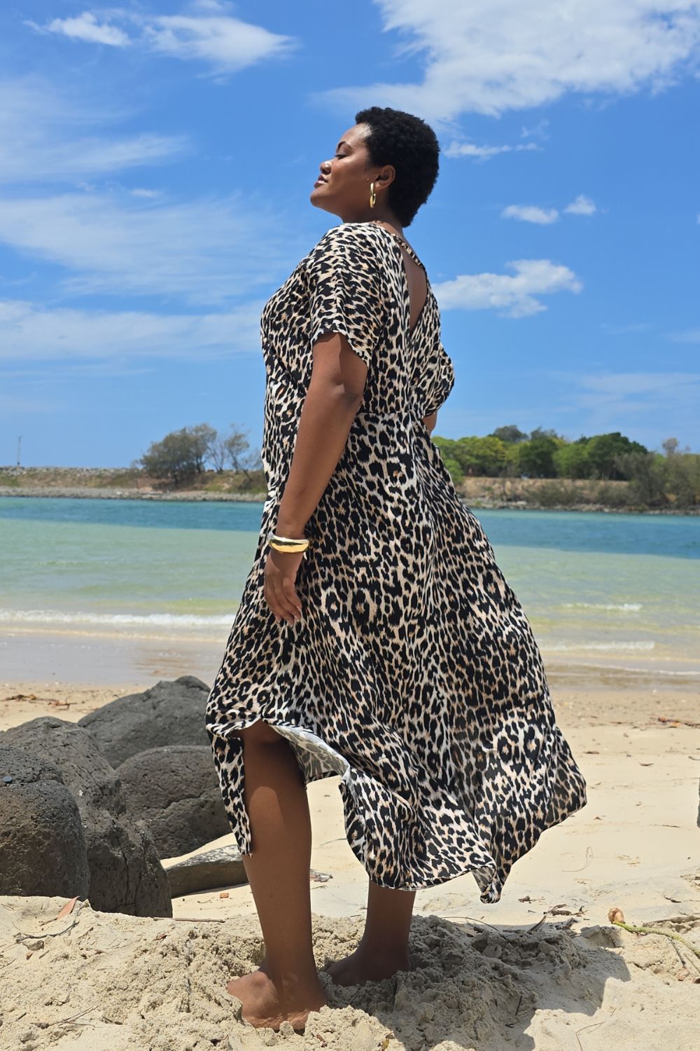 Woman in a leopard print dress standing on a Queensland beach with a blue sky and water in the background