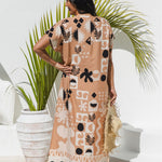A woman stands with her back to the camera wearing a antique brass maxi kaftan dress with a black and ghost white abstract tribal pattern. She is holding a straw hat and standing in front of a white wall with a potted palm plant.