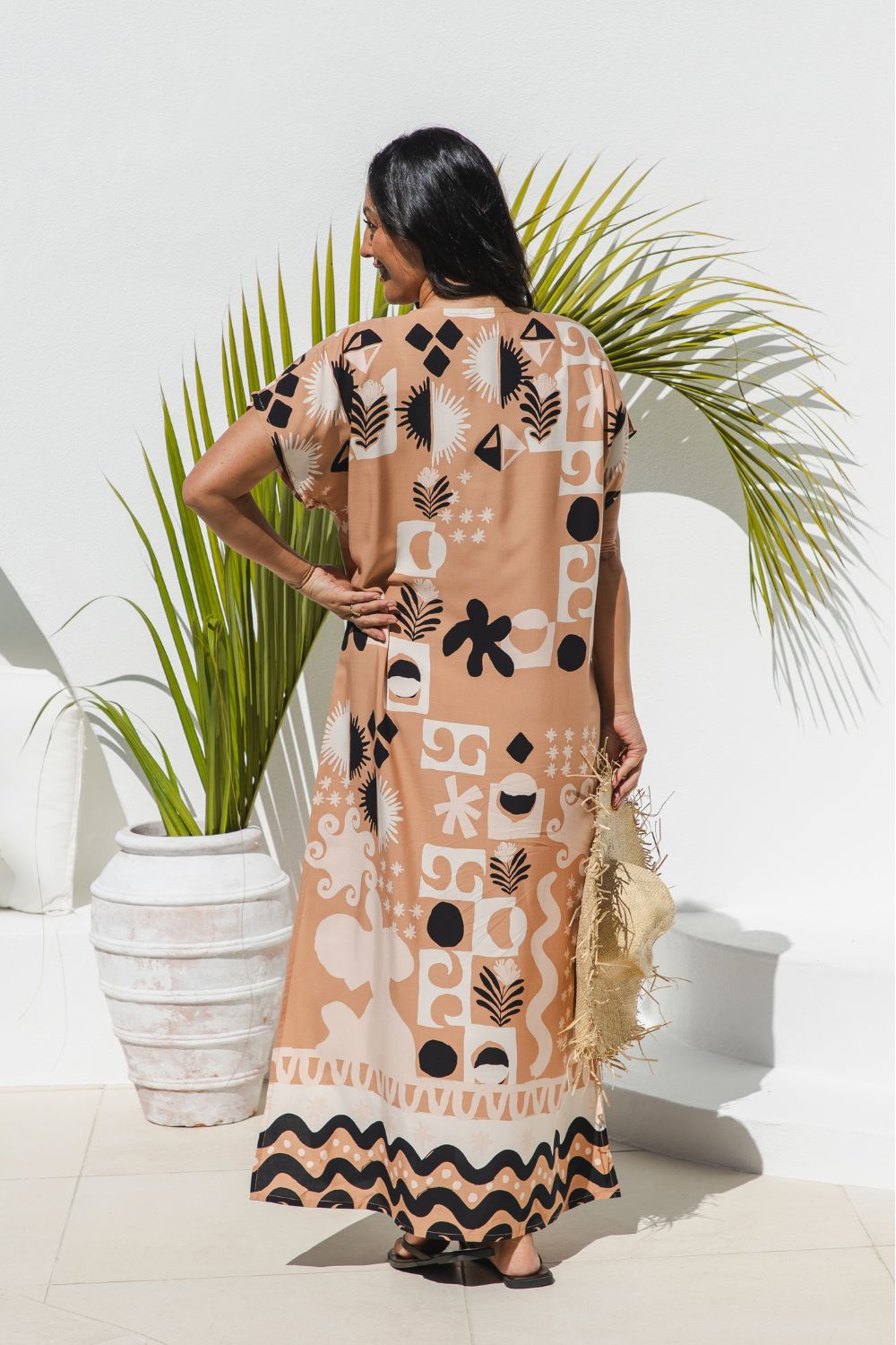 A woman stands with her back to the camera wearing a antique brass maxi kaftan dress with a black and ghost white abstract tribal pattern. She is holding a straw hat and standing in front of a white wall with a potted palm plant.