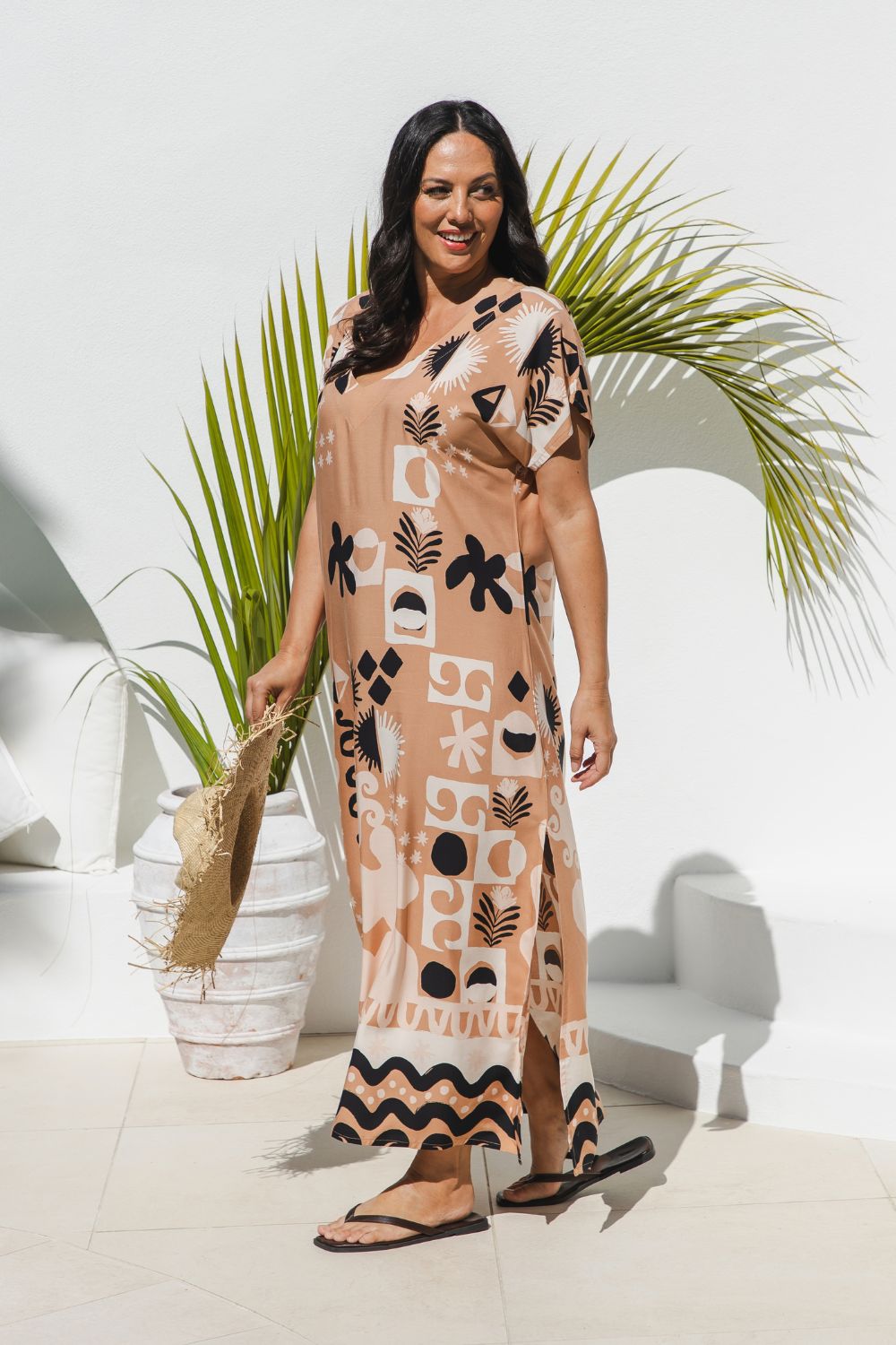 A mature woman modelling a soft terracotta maxi dress with black and ghost white geometric and botanical patterns, standing in a outdoor area poolside with potted palm behind