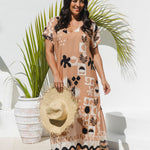 A woman modeling a peach maxi kaftan dress with black and white abstract pattern, holding a straw hat, standing in front of a white wall with a potted palm.