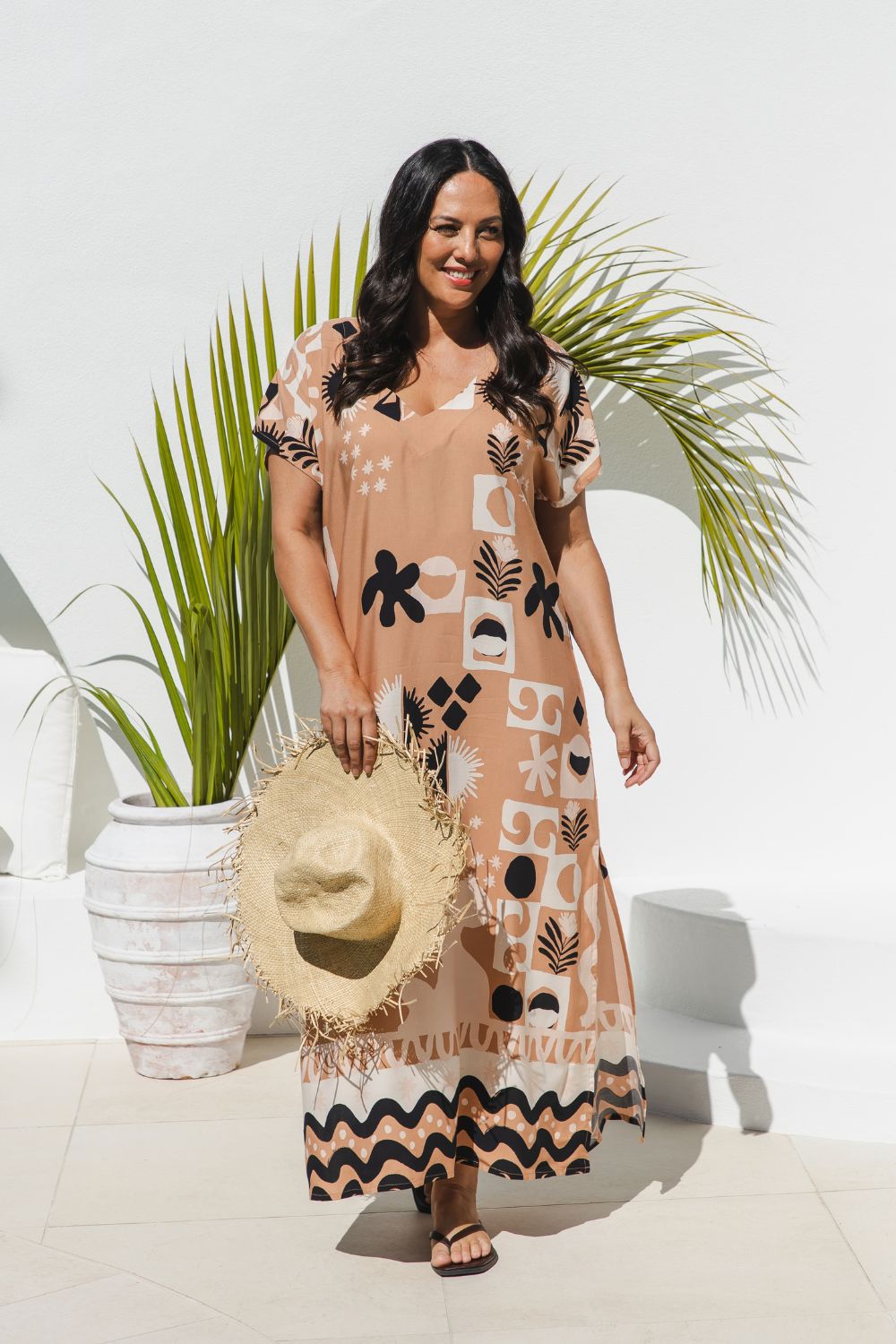 A woman modeling a peach maxi kaftan dress with black and white abstract pattern, holding a straw hat, standing in front of a white wall with a potted palm.