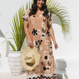 A woman modeling a peach maxi kaftan dress with black and white abstract pattern, holding a straw hat, standing in front of a white wall with a potted palm.