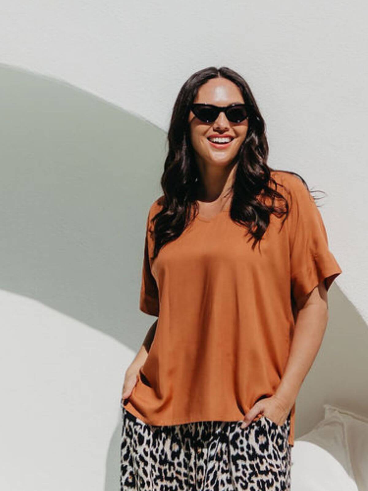 Model wearing mocha mousse blouse top standing in sunlight against white wall with leopard pants