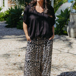A woman wearing a black kimono sleeve kaftan top and leopard-print wide-leg pants, standing outdoors on a gravel path with tropical plants in the background.