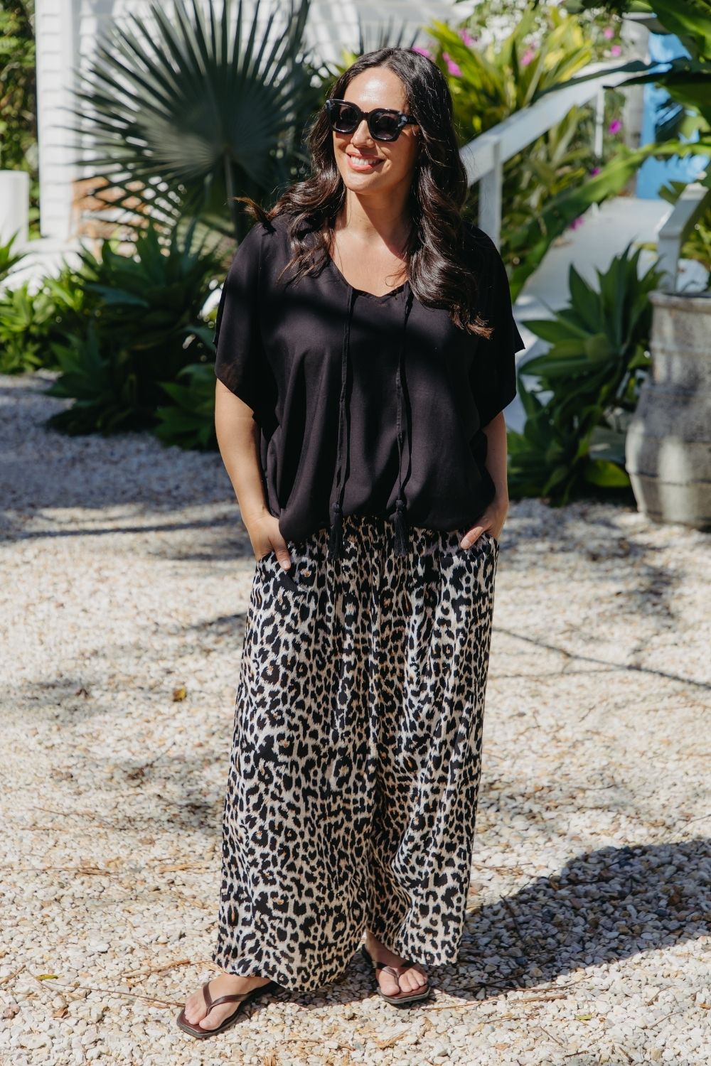 A woman wearing a black kimono sleeve kaftan top and leopard-print wide-leg pants, standing outdoors on a gravel path with tropical plants in the background.