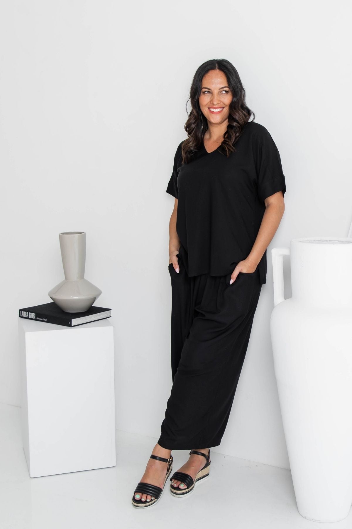 A woman modeling a black short-sleeve V-neck top and matching black wide-leg pants, standing in a bright white studio setting with decorative vases.