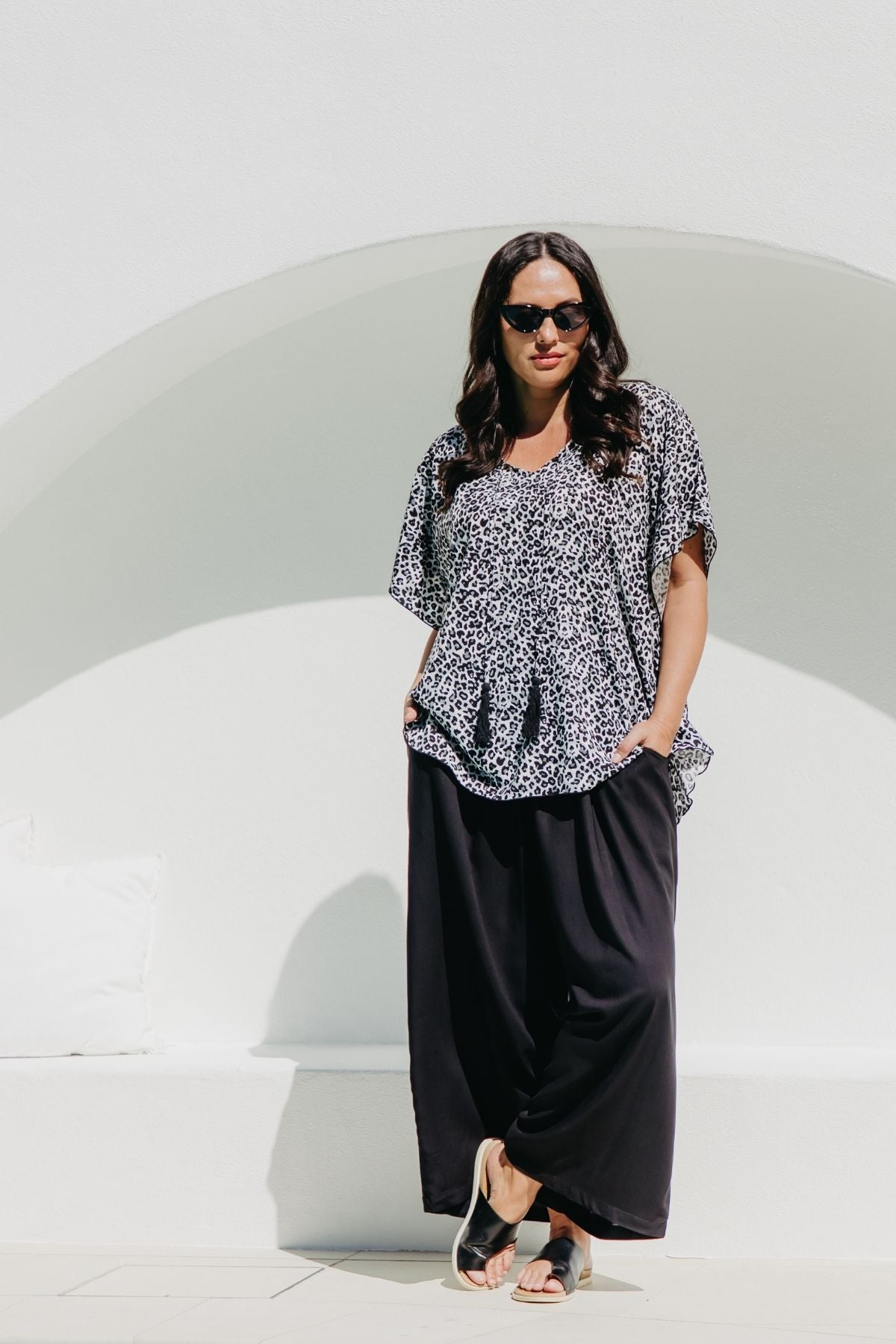 A woman modeling a kimono-sleeve black grey and white leopard print kaftan top with black wide-leg pants and black sandals, standing against arched lounge area near the pool