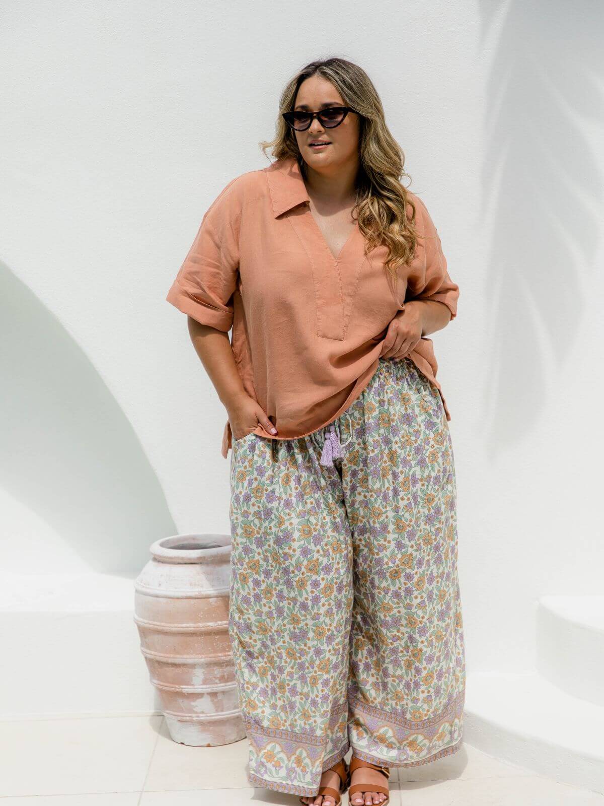 Plus size model wearing floral print palazzo pants against sunlit white wall with palm leaf shadow