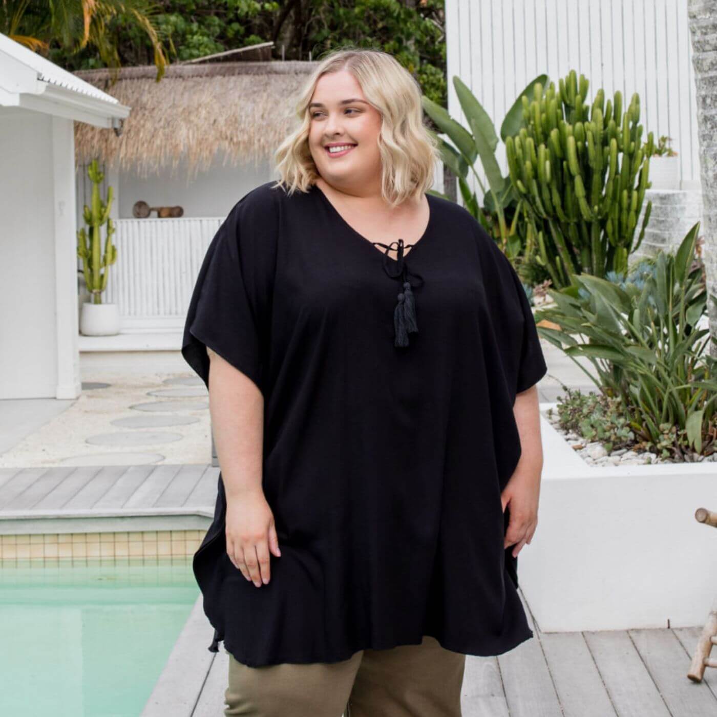 Plus size model wearing a black kaftan top standing by a pool with lush gardens in the background