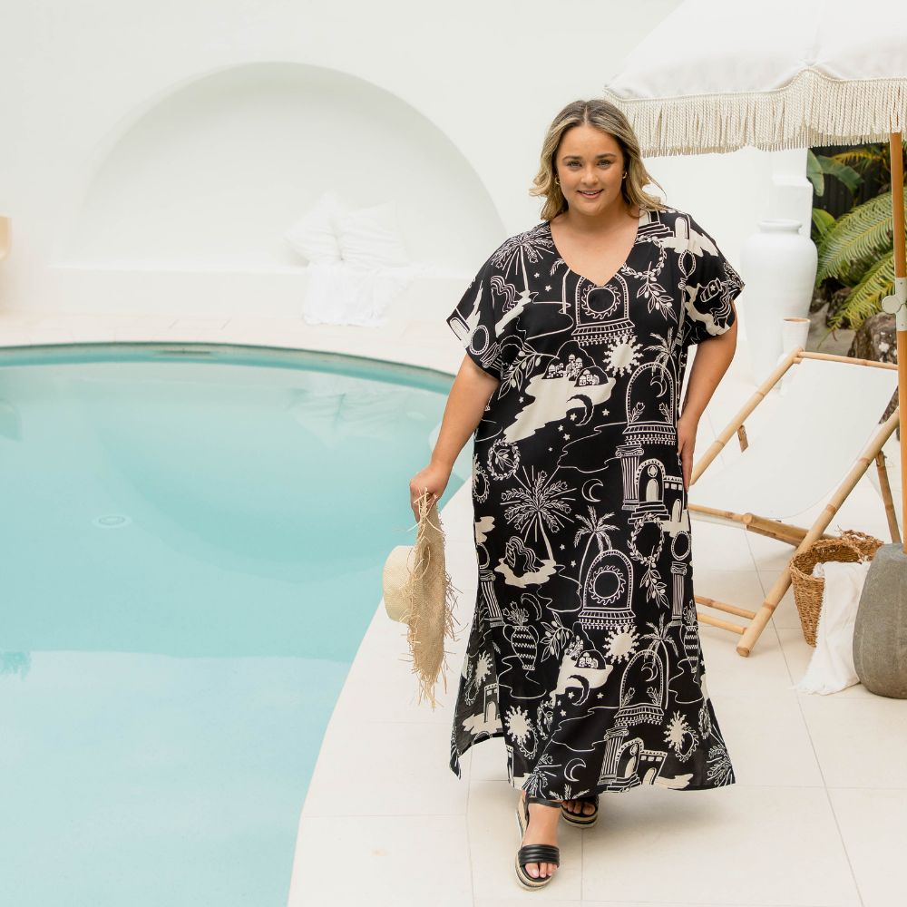Plus size model wearing a black and cream graphic print long kaftan dress standing poolside holding a woven sunhat