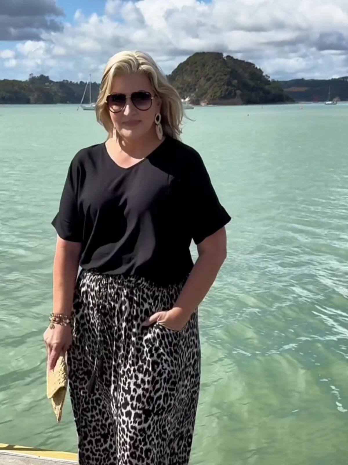 Mature woman wearing a black blouse and leopard print wide leg pants standing on a jetty with the Bay of Islands in the background