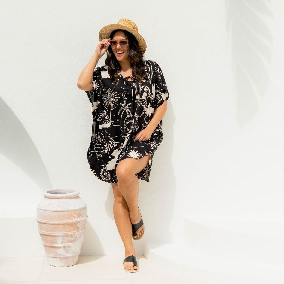 Woman wearing a black and cream graphic print design short kaftan dress and straw hat against a white background