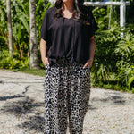 A woman wearing a black kaftan top and relaxed fit leopard-print pants in outdoors tropical greenery setting