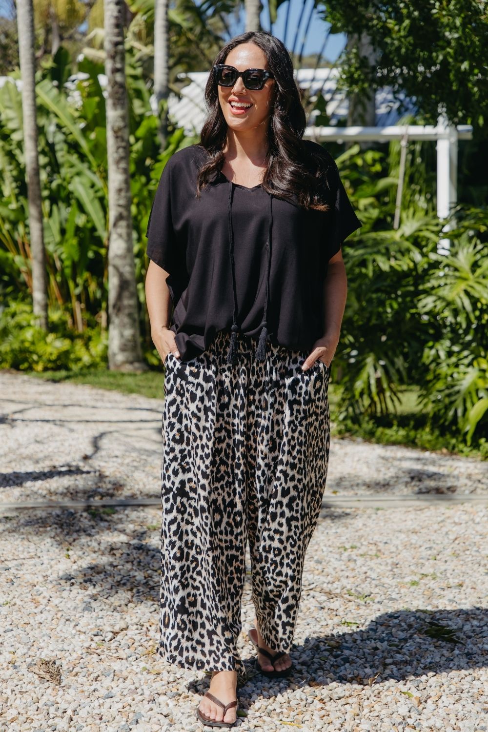 A woman wearing a black kaftan top and relaxed fit leopard-print pants in outdoors tropical greenery setting