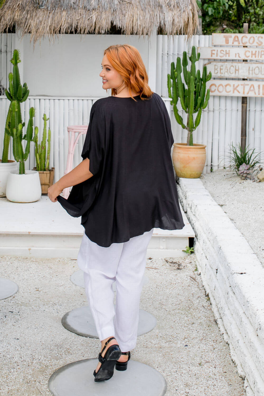 Back view of a woman wearing a black kaftan top walking away, showing length and drape on a size S/M model