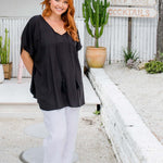 Woman wearing a black kaftan top standing near a beach bar, showing relaxed drape over white pants and tassel detail on a size S/M model
