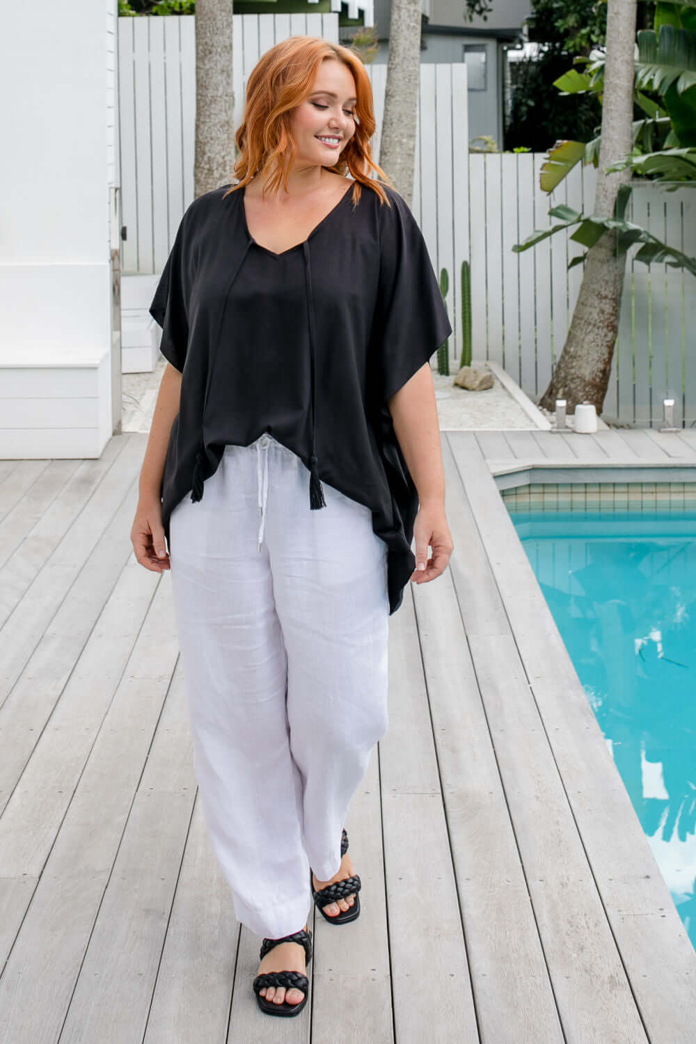 Woman walking poolside wearing a black kaftan top styled with a centre front tuck on a size S/M model