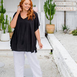 Woman standing near a garden rock wall wearing a black kaftan top over white pants on a size S/M model
